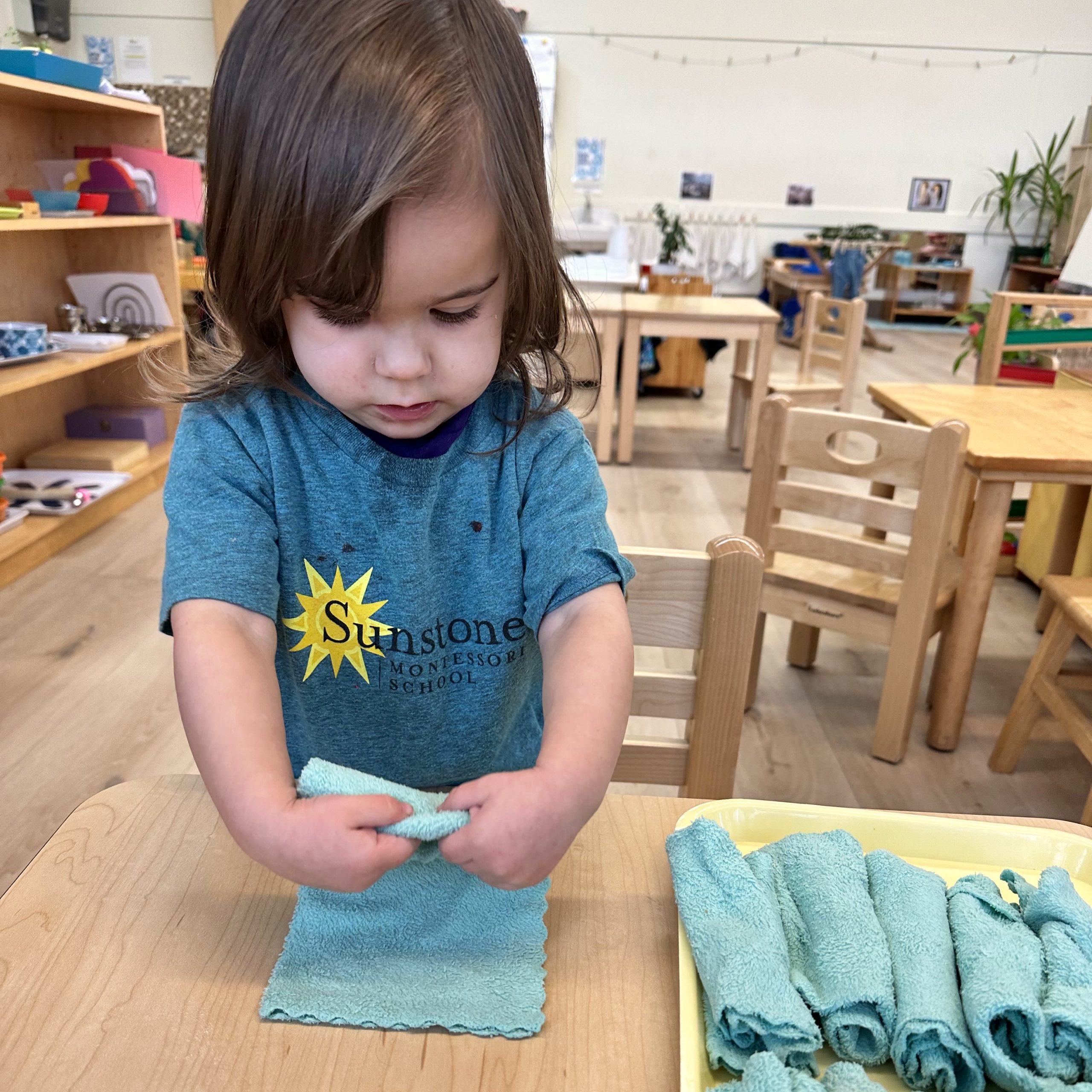 A toddler folds a small cloth at a classroom table