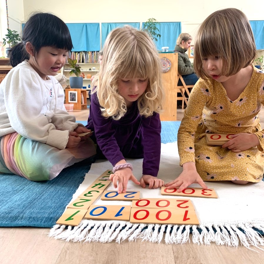 Three young students build numbers with Montessori number cards on a rug