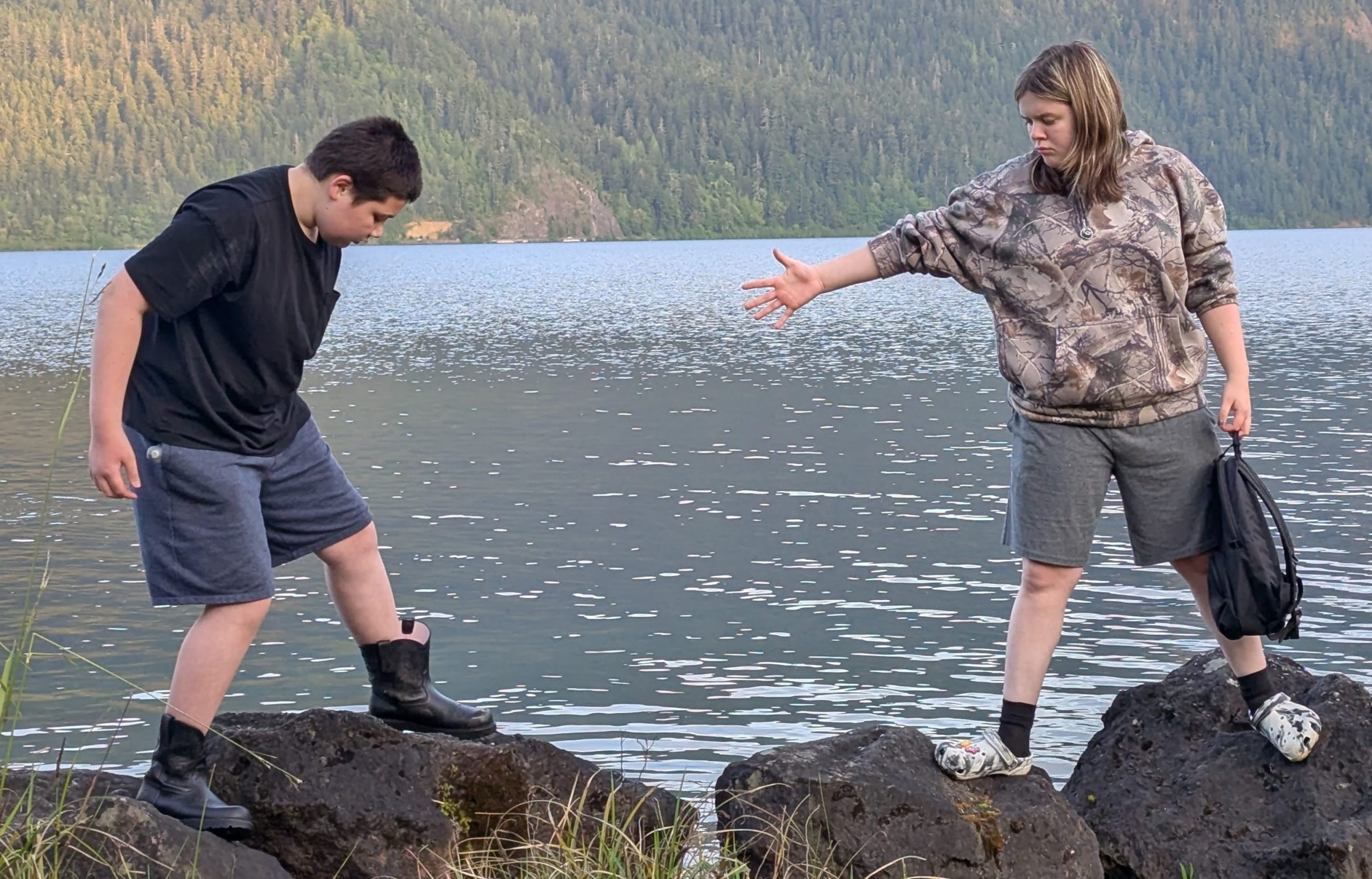 A teacher reaches out to a student stepping across rocks at a lakeshore