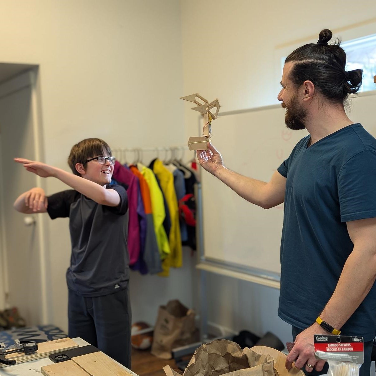 A middle school student gestures while presenting a small wooden model to a smiling teacher