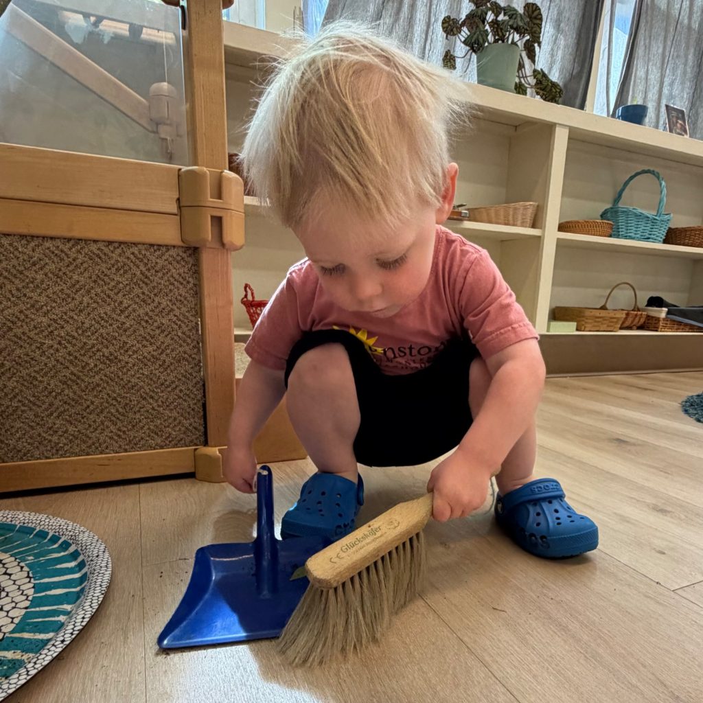 A toddler sweeps the floor with a brush and dustpan