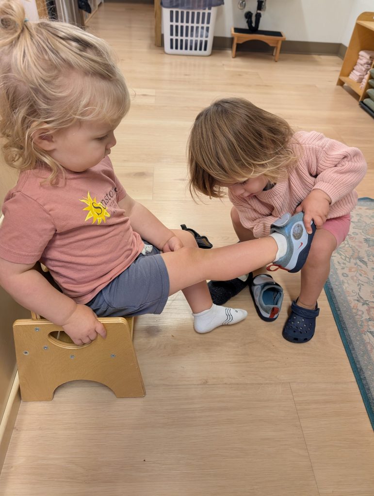 One toddler helps their classmate put a shoe on, mentorship, collaboration, empathy starts early at Sunstone