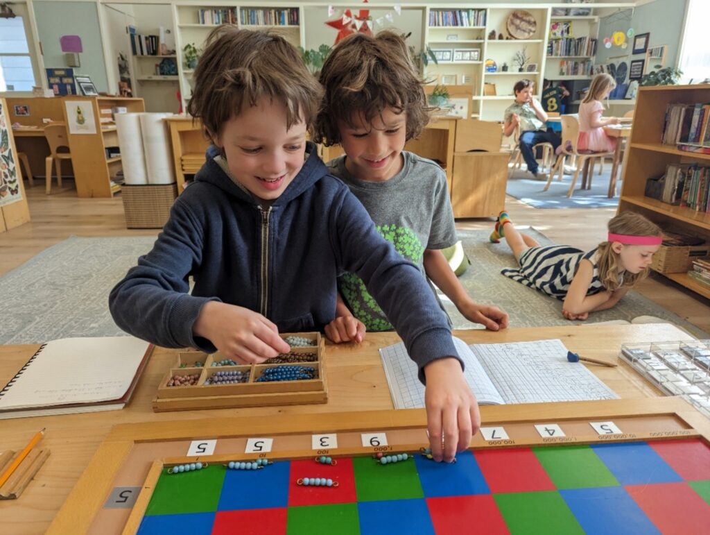 Two boys work the Montessori checkerboard with colored bead bars to solve a large multiplication problem.