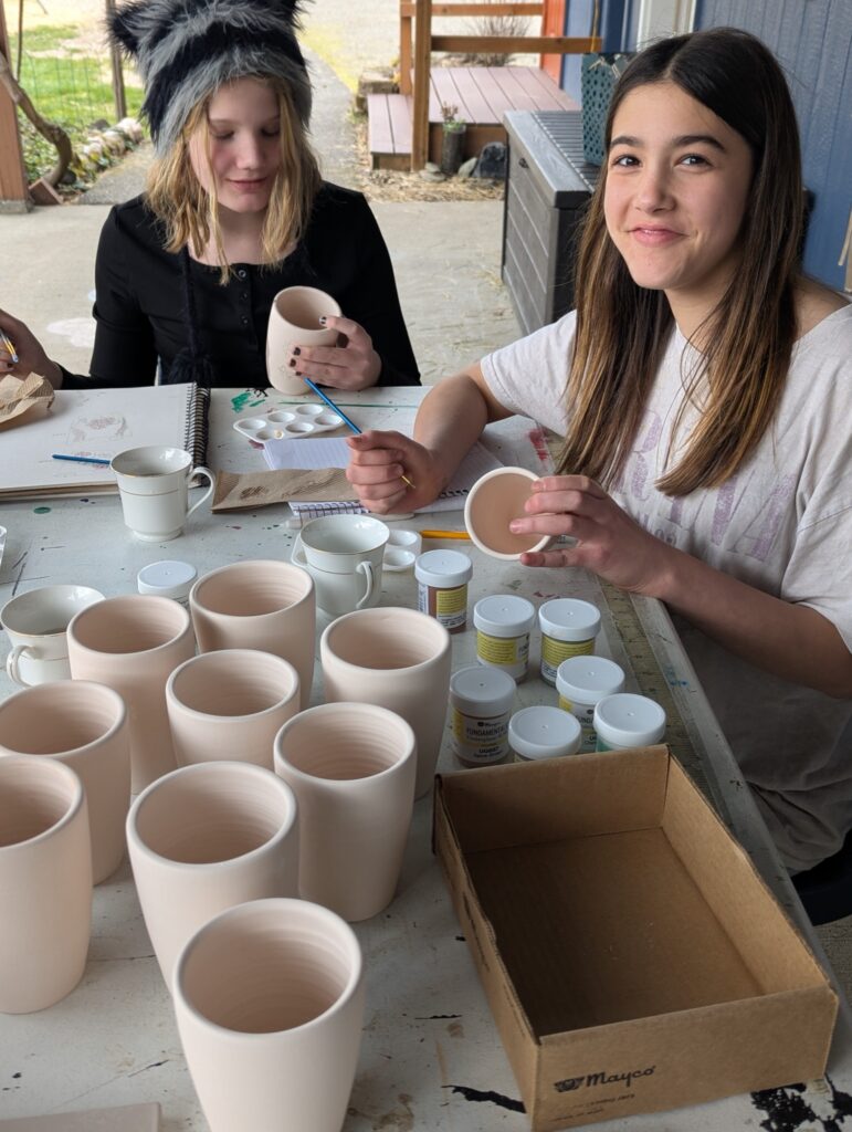 Two students glaze rows of handmade ceramic mugs at an outdoor table, one smiling at the camera.