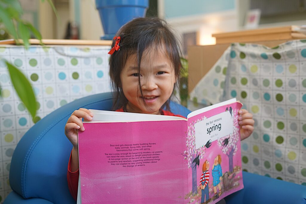 A young child sits in a soft chair smiling while holding up an open picture book about spring.