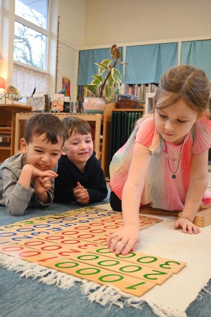 A young child lays out wooden number cards on a mat while two other children watch closely.