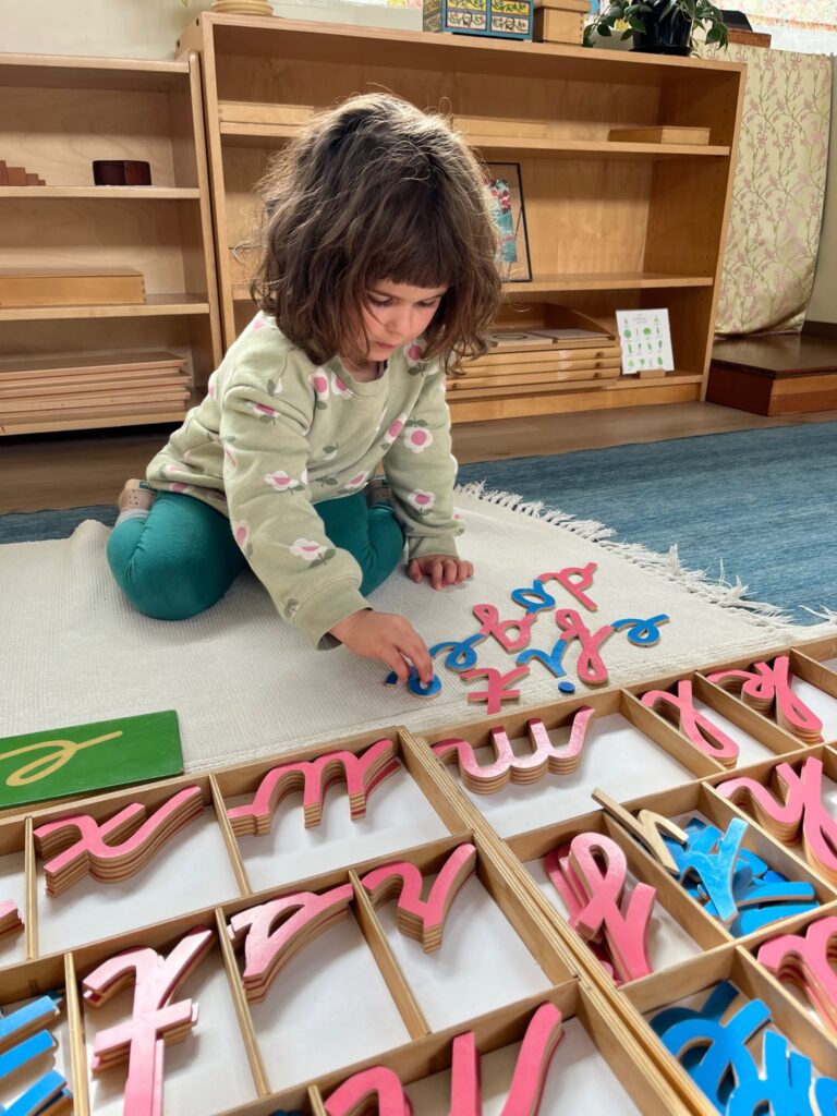 A young child kneels on a mat arranging pink and blue movable alphabet letters from wooden trays.