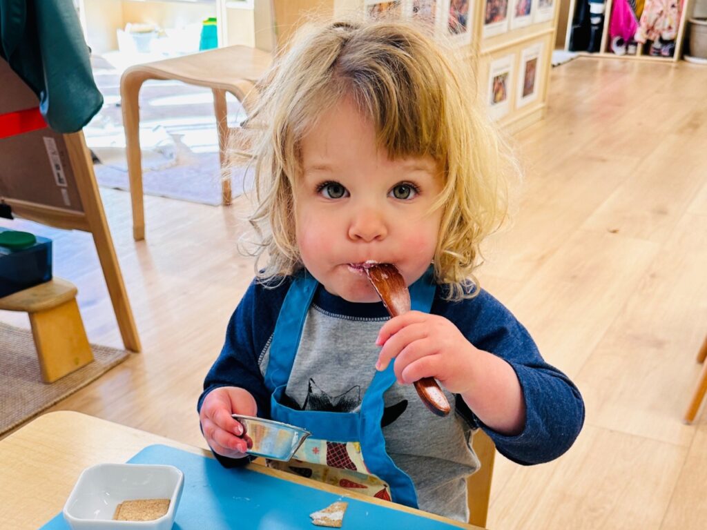 A toddler in an apron tastes from a wooden spoon during a self-serve snack at the table.