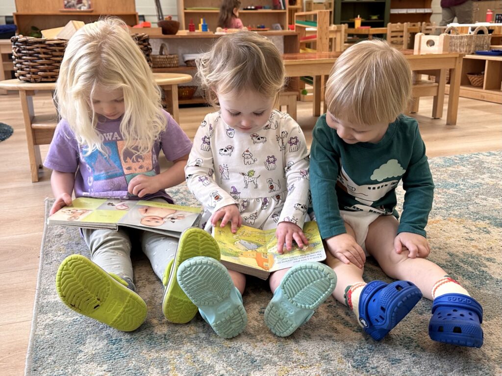 Three toddlers sit on the rug sharing a picture book in a sunny classroom.