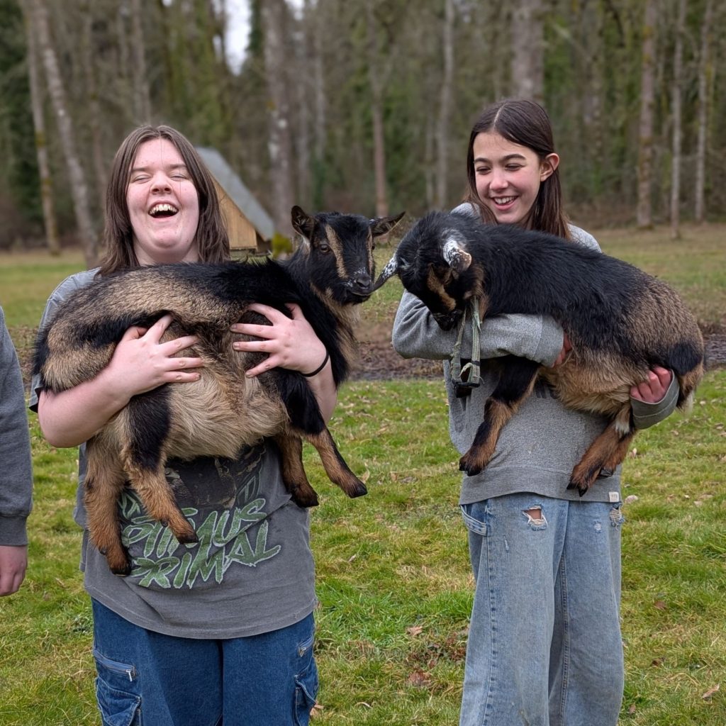 Two middle school students laugh while holding young goats at the farm campus