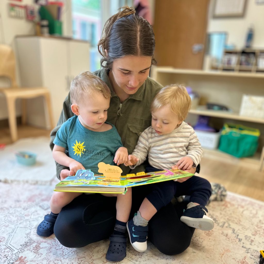a teacher sits cross-legged with one toddler on each knee as they read a book together